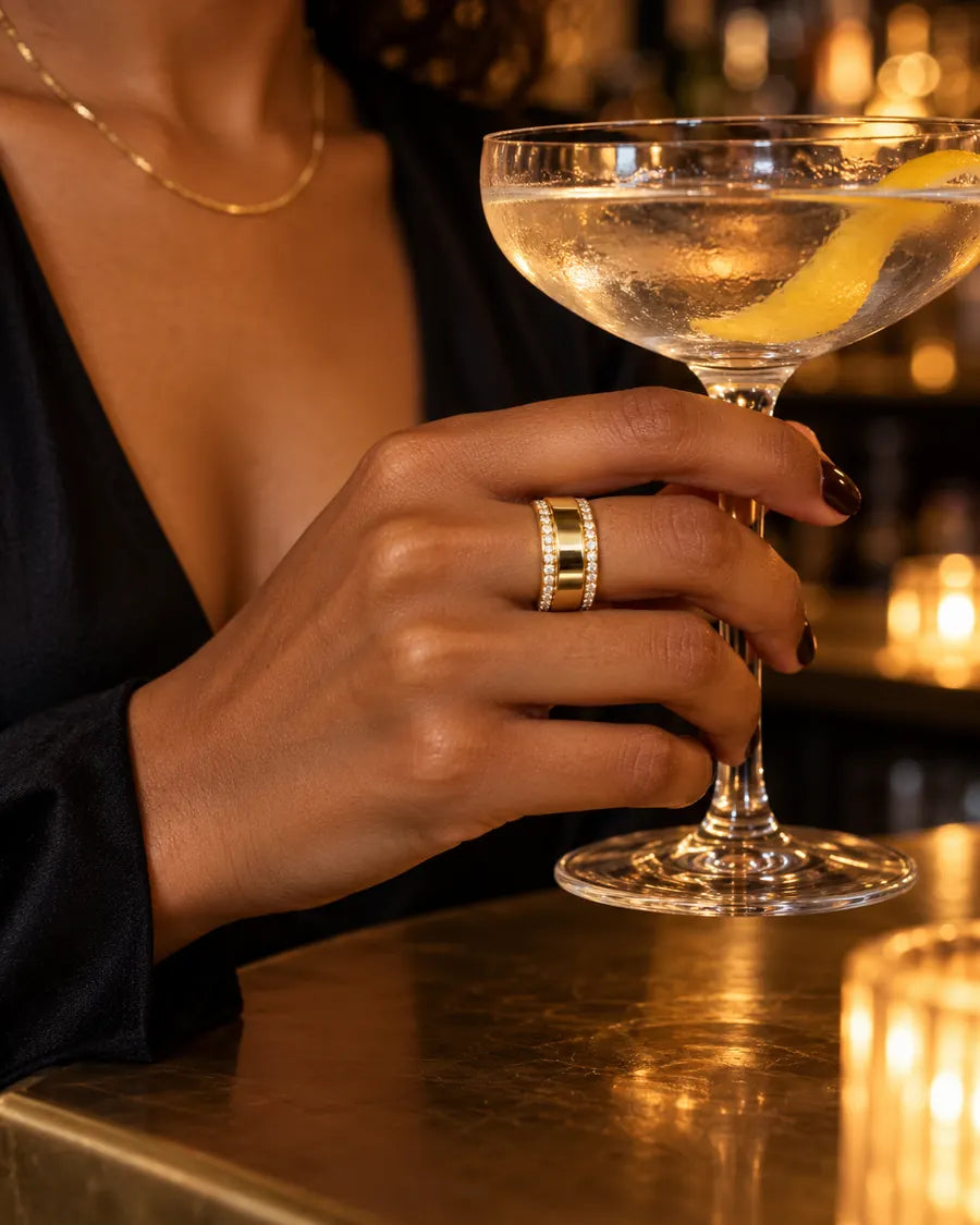HALO Round in 14k yellow gold with round diamonds, worn over an Oura Ring while holding a coupe cocktail at a candlelit bar